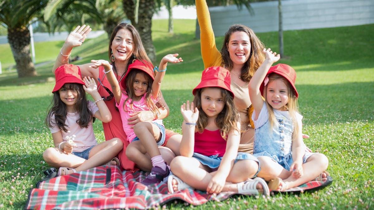 Two women and four girls enjoy a sunny picnic in Portugal, waving happily in the park.